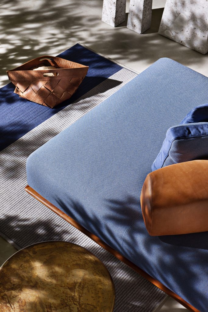 Caprera Island sofa. Upholstered in blue fabric, base in brown leather and blue and brown cushions on a terrace.