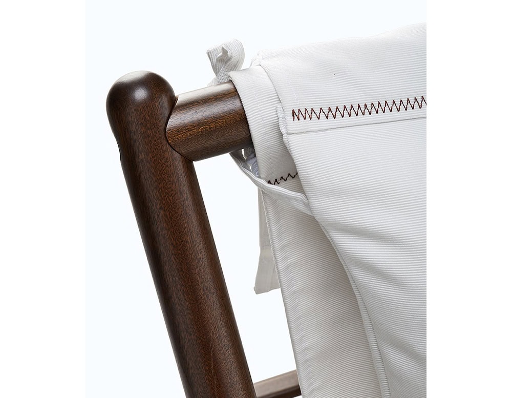 Closeup of Paraggi chair with crossed legs made of brown wood, upholstered in white fabric on a white background.