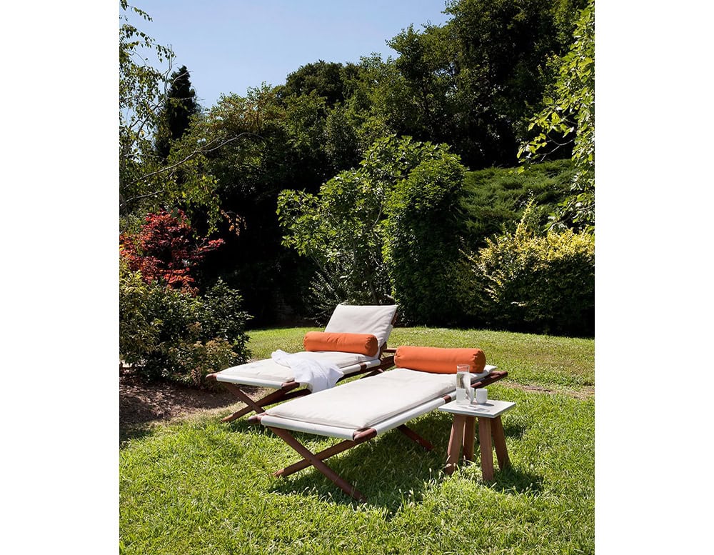 Two Paraggi Sun Loungers in brown wood upholstered in white fabric with orange cushion in a garden.